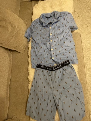 Blå pyjamas från Polo Ralph Lauren - Snygg blå pyjamas från Polo Ralph Lauren med kortärmad skjorta och shorts. Setet har ett klassiskt mönster med små broderade loggor och svarta resårband med varumärkesnamnet på shortsen. Perfekt för chillkvällar hemma.