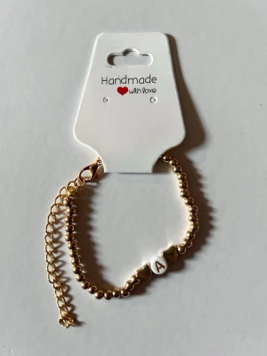 Guldfärgat pärlarmband med bokstav A - Stilrent armband med små guldfärgade pärlor och en vit pärla med bokstaven A. Armbandet har justerbar kedja och hjärtformade detaljer. Perfekt accessoar för att ge en personlig touch till din stil.