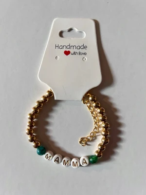 Guldfärgat pärlarmband med text MAMMA - Stilrent armband med guldfärgade pärlor och vita bokstavspärlor som bildar ordet 'MAMMA'. Två gröna pärlor ger extra färg. Justerbar kedja för perfekt passform. Perfekt accessoar för att visa uppskattning till mamma.