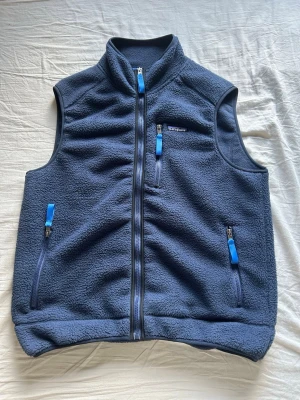 Blå fleeceväst från Patagonia - Mysig blå fleeceväst från Patagonia med hög krage och hel dragkedja. Västen har tre fickor med blå dragkedjor och klassisk logga på bröstet. Perfekt för lager-på-lager och outdoor vibes.