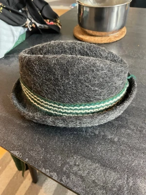 Grå filthatt med randigt band one size - Säljer en grå filthatt med coolt grönt och vitt randigt band runtom. Hatten har en klassisk form med uppvikt brätte och är tillverkad i grov filt. Perfekt för dig som vill sticka ut med en unik accessoar.