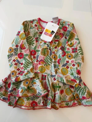 Blommig klänning från Re-Kid - Färgglad blommig klänning från Re-Kid med lång ärm och volang nertill. Klänningen har en vit bas med mönster i grönt, rosa, gult och rött samt rund halsringning med rosa kant. Perfekt för dig som gillar lekfull stil!