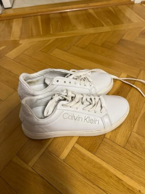 Vita Calvin Klein sneakers - Snygga vita sneakers från Calvin Klein med klassisk låg profil och logga i silver på sidan. Skorna har vita snören och ovandel i slätt läderliknande material. Perfekta för en clean och stilren look.
