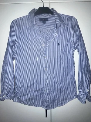 Blåvit randig skjorta från Ralph Lauren - Klassisk blå och vit randig skjorta från Ralph Lauren med lång ärm och knappar framtill. Skjortan har en broderad logga på bröstet och är tillverkad i bomull. Perfekt för en stilren och avslappnad look.