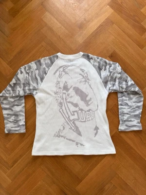 Vit långärmad thermal topp med camoärmar - Cool långärmad topp från Sangue di Lupo med vita och grå camouflagemönstrade ärmar. Framsidan är enkel medan baksidan har ett stort tryck av en varg och Lupo-loggan. Tillverkad i ett strukturerat (thermal) bomullsmaterial som känns sportigt och bekvämt.