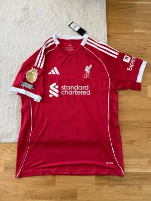 Liverpool matchtröja Adidas röd - Röd Liverpool FC matchtröja från Adidas med vita detaljer och klubbmärke på bröstet. Tröjan har Premier League-märke på ärmen, sponsorloggor och klassiska Adidas-ränder på axlarna. Perfekt för fotbollsfans som vill visa sitt stöd på läktaren eller planen.