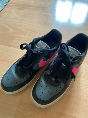 Nike Air Force 1 svart med rosa swoosh - Nike Air Force 1 sneakers i svart läder med en iögonfallande rosa swoosh på sidorna. Klassisk siluett med perforerad tå, svarta skosnören och vit yttersula.                     Innermått 24,5 cm