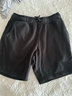 Svarta mjukisshorts med snörning - Svarta shorts i mjukt material med resår och snörning i midjan. De har två sidofickor och en avslappnad passform, perfekta för chill dagar eller träning.