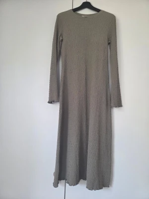 Beige långärmad långklänning - En stilren beige långklänning med lång ärm och rund halsringning. Klänningen har en lätt strukturerad yta och ett rakt, flödande fall som ger en avslappnad men elegant look. Perfekt för dig som gillar minimalistisk stil.