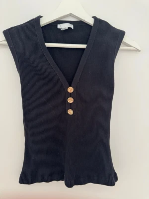 Svart ribbad ärmlös topp med knappar - Snygg svart ribbad topp med djup V-ringning och tre dekorativa guldknappar framtill. Ärmlös modell i stretchigt material som sitter tajt och passar perfekt till jeans eller kjol. Enkel men trendig design som är lätt att matcha.