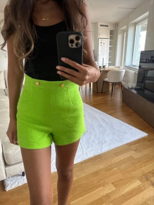 Limegröna högmidjade shorts med guldknappar från Zara - Snygga limegröna högmidjade shorts med två guldknappar framtill. Shortsen har en strukturerad yta och är perfekta för en trendig look. Passformen är normal och materialet känns som en blandning av bomull och syntet.