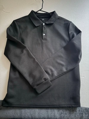 Svart långärmad henley från Grandiety - Säljer en svart långärmad henley från Grandiety med diskret mönstrad struktur. Skjortan har klassisk krage, två vita knappar framtill och är tillverkad i ett mjukt syntetmaterial. Perfekt för dig som gillar stilrena plagg med en twist.