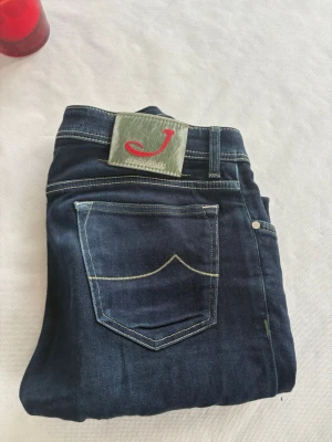 Mörkblå jeans från Jacob Cohen - Snygga mörkblå jeans från Jacob Cohen med klassisk femficksdesign och kontrastsömmar. Bakfickan har dekorativ söm och bak finns en grön patch med röd J-logga. Perfekta för en trendig och avslappnad look.