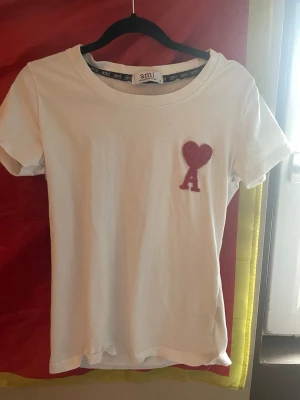 Vit t-shirt från Ami Paris med hjärtdetalj - Vit t-shirt från Ami Paris med klassisk passform och korta ärmar. På bröstet finns en röd hjärtdetalj med bokstaven A. Tillverkad i mjuk bomull och har rund halsringning. Perfekt för en clean och stilren look.