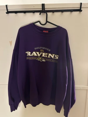 Lila Baltimore Ravens NFL sweatshirt - Säljer en lila oversized sweatshirt med Baltimore Ravens-tryck på bröstet. Tröjan har rund halsringning och ribbade muddar vid ärmslut och nederkant. Perfekt för dig som gillar amerikansk fotboll och streetwear.
