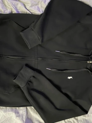 Svart hoodie från Polo Ralph Lauren - Svart hoodie med dragkedja från Polo Ralph Lauren. Klassisk design med vit broderad logga på bröstet, huva med dragsko och ribbade muddar. Perfekt för en chill och stilren look. Materialet är mjukt och bekvämt, troligtvis bomullsblandning. Storlek s men sitter tajt i midjan