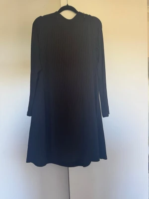 Svart ribbad klänning från Indiska - Svart långärmad klänning från Indiska i ribbad struktur. Klänningen har en enkel, rak passform och är tillverkad i ett mjukt material som faller snyggt. Perfekt för dig som gillar stilrena och bekväma plagg.