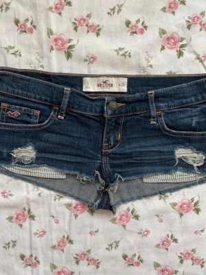 Mörkblå jeansshorts från Hollister - Skitsnygga super low waist mörkblå jeansshorts från hollister med slitna detaljer och råa kanter. Klassisk femficksmodell med ultra låg midja och logga på fickan. Perfekta för varma dagar och enkel att matcha med allt. Midjemått: 38cm