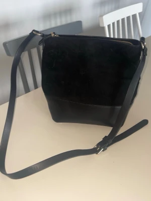 Svart axelväska i mocka och läder - Snygg svart axelväska med justerbar rem. Väskan är tillverkad i en kombination av mocka och slätt läder, vilket ger en stilren och modern look. Stängs med dragkedja upptill och har en rymlig form som passar till mycket.