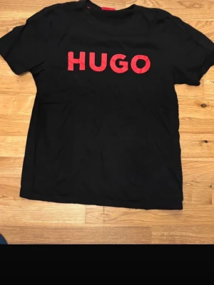 Svart HUGO t-shirt med röd logga - Svart t-shirt från HUGO med stor röd logotyp framtill. Klassisk rund halsringning och korta ärmar. Perfekt för dig som gillar stilrena plagg med tydlig branding. Tillverkad i mjuk bomull för skön känsla hela dagen.