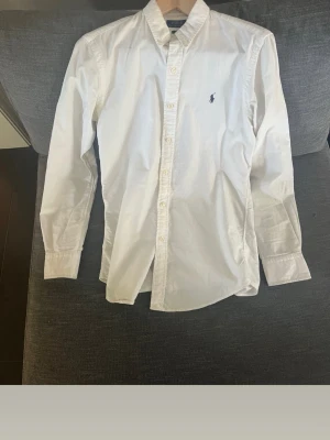 Vit skjorta från Polo Ralph Lauren - Klassisk vit skjorta från Polo Ralph Lauren med lång ärm och knappar framtill. Skjortan har en stilren passform och ikonisk broderad logga på bröstet. Perfekt till jeans eller chinos för en clean look. Smutsig och fläckig 