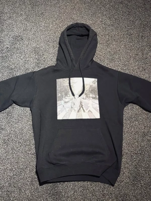 Svart hoodie med Abbey Road-tryck - Svart hoodie med klassiskt Abbey Road-motiv på bröstet. Hoodien har huva med dragsko och en stor magficka. Perfekt för dig som gillar musik och vill ha en stilren streetwear-look.