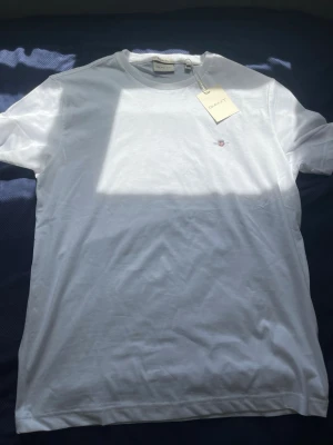 Vit t-shirt från Gant i bomull - Klassisk vit t-shirt från Gant med liten broderad logga på bröstet. T-shirten har rund halsringning och är tillverkad i mjuk bomull. Perfekt basplagg med enkel och stilren design.