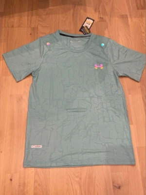 Ljusgrön träningströja från Under Armour - Snygg ljusgrön t-shirt från Under Armour med diskret mönster och färgglad logga på bröstet. Tillverkad i ett lätt och snabbtorkande funktionsmaterial, perfekt för träning. Rund halsringning och korta ärmar.