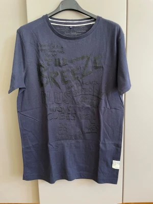 Mörkblå t-shirt med tryck - Snygg mörkblå t-shirt med svart texttryck på framsidan. T-shirten har rund halsringning med dubbla vita ränder på insidan av kragen och korta ärmar. Perfekt för en avslappnad och trendig stil.