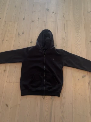 Svart hoodie från Polo Ralph Lauren - Svart hoodie med dragkedja från Polo Ralph Lauren. Klassisk design med huva, känguruficka och den ikoniska vita logon broderad på bröstet. Perfekt för en avslappnad och stilren look. Mjuk bomullsblandning för extra komfort.