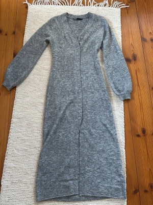 Grå ribbad långklänning Bik Bok - Grå långklänning från Bik Bok i ribbat material med v-ringning och långa ärmar. Klänningen har knappar hela vägen framtill och är figurnära med en mjuk och stretchig känsla. Perfekt för dig som gillar enkel och stilren design.
