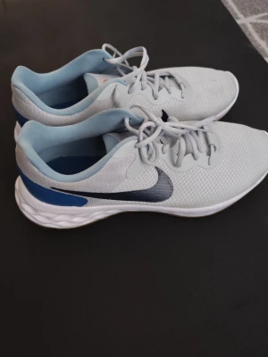 Nike vita och blå sneakers - Snygga sneakers från Nike i vitt meshmaterial med ljusblå detaljer och mörkblå swoosh på sidan. Skorna har en låg siluett, vita snören och en bekväm, vadderad sula. Perfekta för en sportig och avslappnad stil.