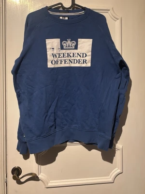 Blå Weekend Offender sweatshirt - Blå sweatshirt från Weekend Offender med vit tryckt logga och text på bröstet. Tröjan har rund halsringning, långa ärmar och ribbade muddar vid ärmslut och nederkant. Perfekt för en avslappnad streetwear-look. Trycket e värme skadat då den låg vikt i solen o trycket smälte men inga andra defekter. Passar S men står M