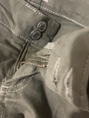 Olivgröna jeans med vita zigzag sömmar Nelly  - Snygga olivgröna jeans med utsvängda ben och vit design på bakfickorna. Jeansen är lågmidjade och väldigt långa. Bakfickorna har zigzag sömmar. 