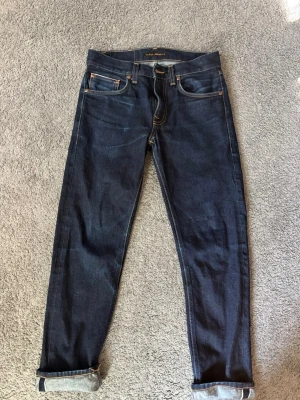 Selvedge Nudie Jeans - Säljer ett par mörkblå jeans från Nudie Jeans med klassisk femficksmodell och kontrastsömmar. Jeansen har upprullade benslut och är tillverkade i kraftig denim. Passformen är smal och modellen är tidlös, perfekt till en avslappnad stil.