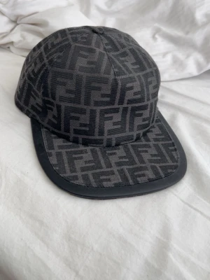 Svart keps med Fendi-logga - Snygg svart keps från Fendi med ikoniskt FF-mönster över hela kepsen. Kepsen har böjd skärm med svart kant och är tillverkad i tyg med detaljer i läder. Perfekt för dig som vill ha en exklusiv accessoar med streetkänsla.
