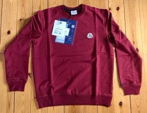 Vinröd sweatshirt från Moncler - Säljer en vinröd sweatshirt från Moncler med rund halsringning och ribbade muddar. Tröjan har Moncler-logga broderad på bröstet och är tillverkad i mjukt bomullsmaterial. Perfekt för en avslappnad och stilren look.