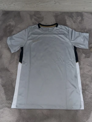 Grå Nike t-shirt med vita detaljer - Säljer en sportig grå t-shirt från Nike med vita paneler på sidorna och svarta inslag vid halsen. Materialet är lätt och andas, perfekt för träning eller sport. Diskret Nike-logga på bröstet och korta ärmar för maximal rörelsefrihet.