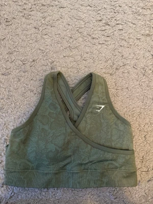 Grön sport-bh från Gymshark - Säljer en grön sport-bh från Gymshark med snyggt mönster och korsade axelband i ryggen. Materialet är stretchigt och bekvämt, perfekt för träning och aktivitet. Logotypen är diskret placerad på framsidan. Passar dig som vill ha både stil och funktion.