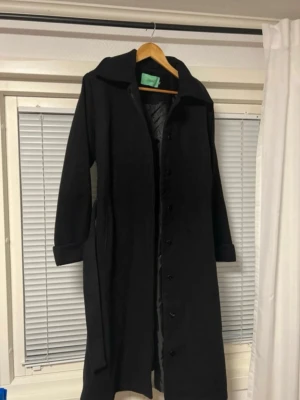 Svart lång rock med bälte - Stilren svart lång rock från Samsøe Samsøe med klassisk krage, knäppning framtill och tillhörande bälte. Rocken har långa ärmar och är fodrad för extra värme. Perfekt för kalla dagar och ger en snygg siluett.