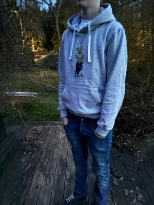 Grå Ralph lauren hoodie - Säljer en grå hoodie från Polo Ralph Lauren med klassiskt björntryck på framsidan. Hoodien har en stor huva med vita snören och en praktisk magficka. Materialet är mjukt och bekvämt.