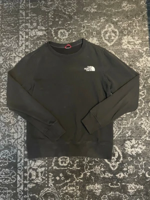 Svart sweatshirt från The North Face - En svart sweatshirt från The North Face med rund hals och logotyp tryckt på bröstet och ryggen. Tröjan har långa ärmar och ribbade muddar vid ärmslut och nederkant. Perfekt för en avslappnad look.