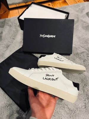 Saint Laurent vita sneakers - Vita sneakers från Saint Laurent med svart detalj på hälen och handskriven logga på sidan. Klassisk låg modell med vit sula och ovandel i slätt läder. Snygg och stilren design som passar till det mesta. Skorna levereras med originalkartong och dustbag.