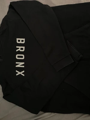 Svart hoodie med BRONX-tryck - Säljer en svart hoodie med stort vitt BRONX-tryck på framsidan. Hoodien har huva, magficka och ribbade muddar vid ärmsluten. Perfekt för en avslappnad streetwear-look.