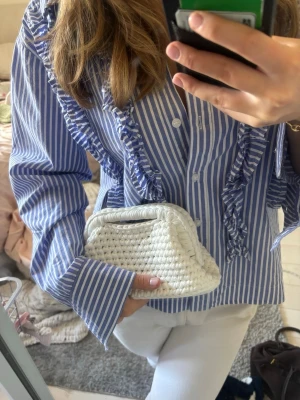 Vit flätad clutchväska - Säljer en vit clutchväska med flätad design och mjukt material. Väskan har ett kompakt format och ett handtag upptill som gör den enkel att bära. Perfekt för att ha med sig det viktigaste och passar till många olika outfits.