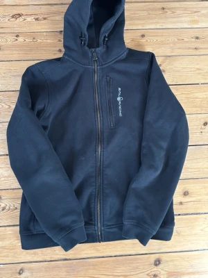 Mörk blå hoodie från Sail Racing - Mörk blå hoodie med dragkedja från Sail Racing. Den har en huva med snörning, ribbade muddar och en bröstficka med dragkedja. Logotypen är tryckt på bröstet. Perfekt för dig som gillar sportig och clean stil.