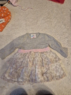 Barn kläder i olika storlekar  - Supersöt klänning från Tiny One med grå överdel i mjuk bomull och långärmad design. Kjolen har två lager med volanger och ett blommigt mönster i ljusa toner, samt ett rosa band i midjan. Perfekt för dig som gillar romantiska detaljer.