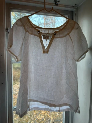 Vit luftig blus med korta ärmar - Säljer en vit, transparent och luftig blus med korta ärmar och rundad halsringning. Blusen har dekorativa knappar och snörning framtill samt beige detaljer längs hals och nederkant. Perfekt för en avslappnad och stilren look.