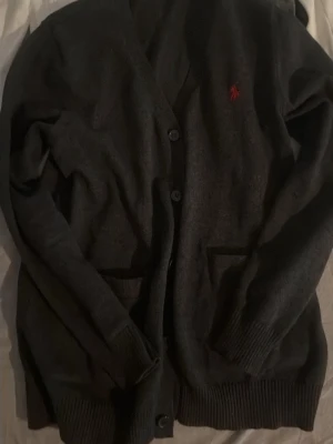 Grå cardigan från Polo Ralph Lauren - Grå cardigan med knappar och två fickor framtill från Polo Ralph Lauren. Klassisk design med ribbade muddar och röd broderad logotyp på bröstet. Perfekt för lager-på-lager och snygg till jeans eller chinos.