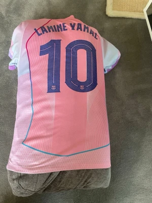 Rosa FC Barcelona matchtröja Nike - Säljer en rosa FC Barcelona matchtröja med Lamine Yamal och nummer 10 på ryggen. Tröjan har blå detaljer, klubbmärke och Nike-logga på framsidan samt Spotify som sponsor. Materialet är lätt och andas, perfekt för fotboll eller träning.